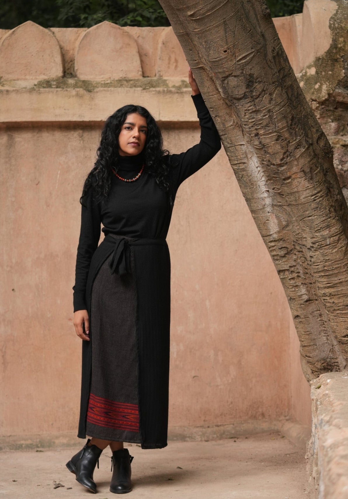 Woman in a black outfit standing against a textured wall with a tree.