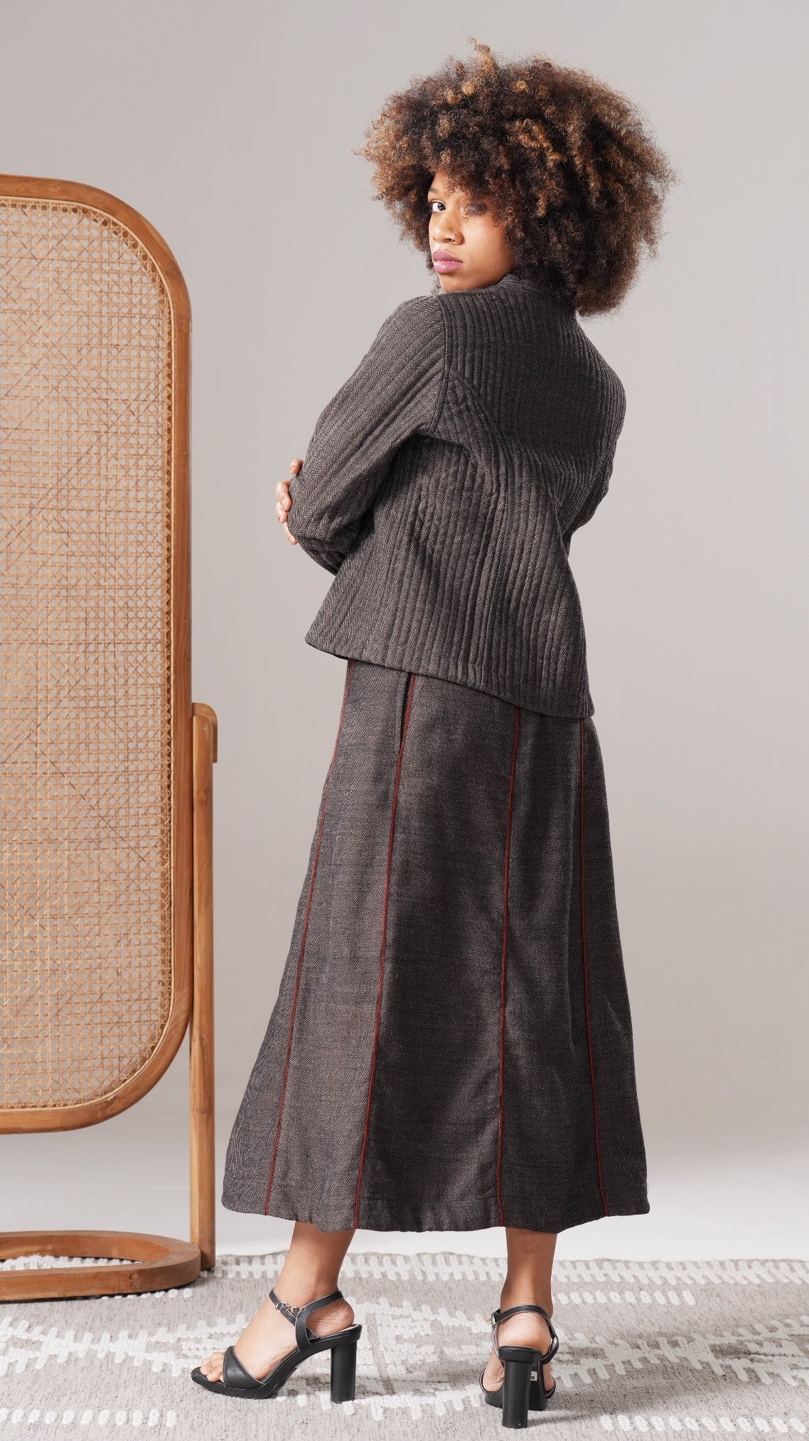 Woman wearing a dark ribbed sweater and patterned skirt standing against a neutral background.