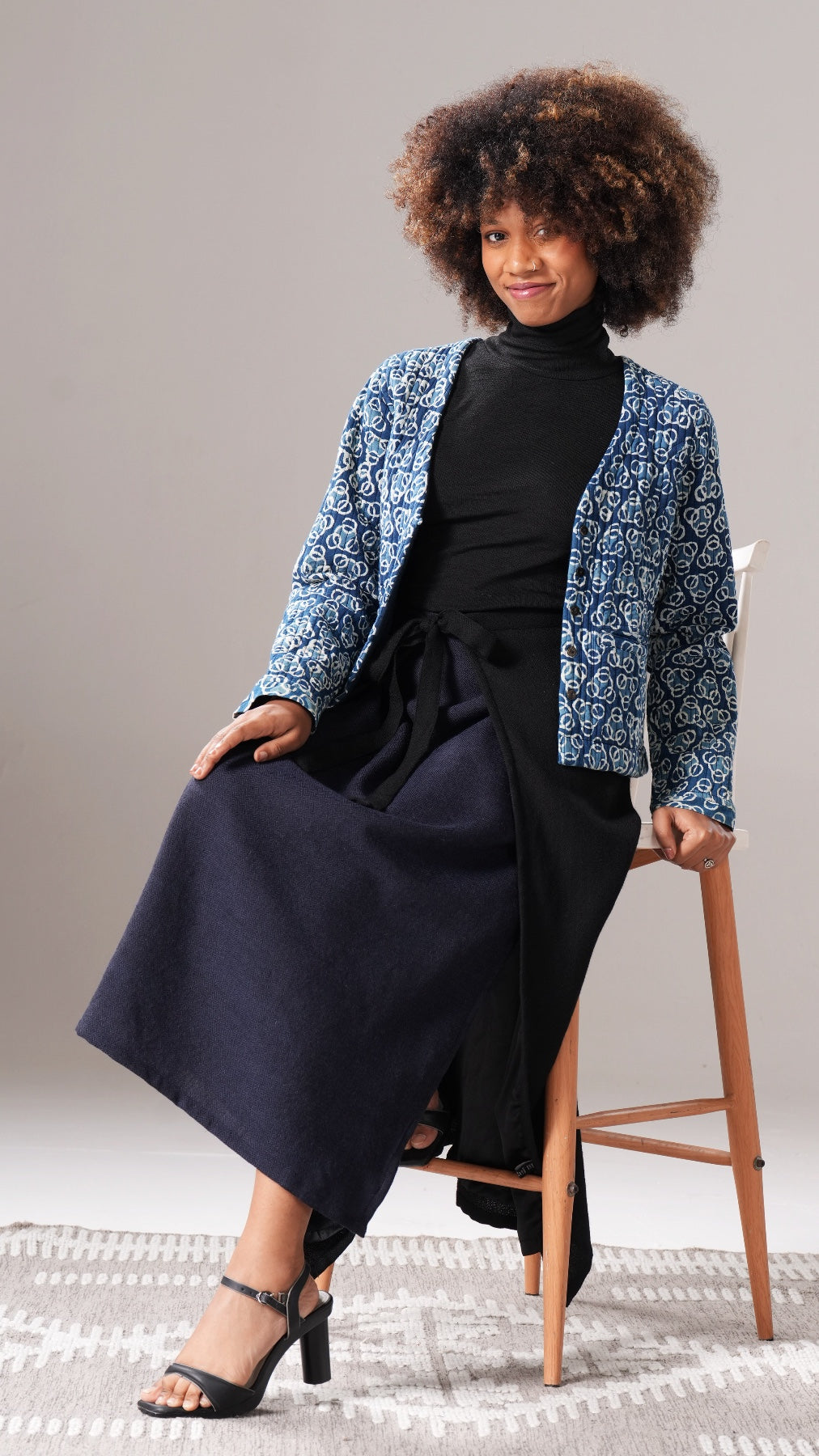 Woman sitting on a stool wearing a black dress and blue patterned jacket against a neutral background