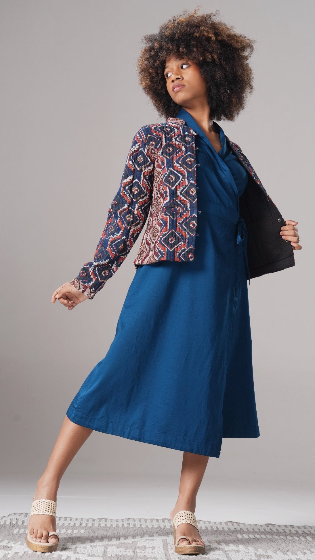 Woman wearing a blue dress and patterned jacket on a plain background