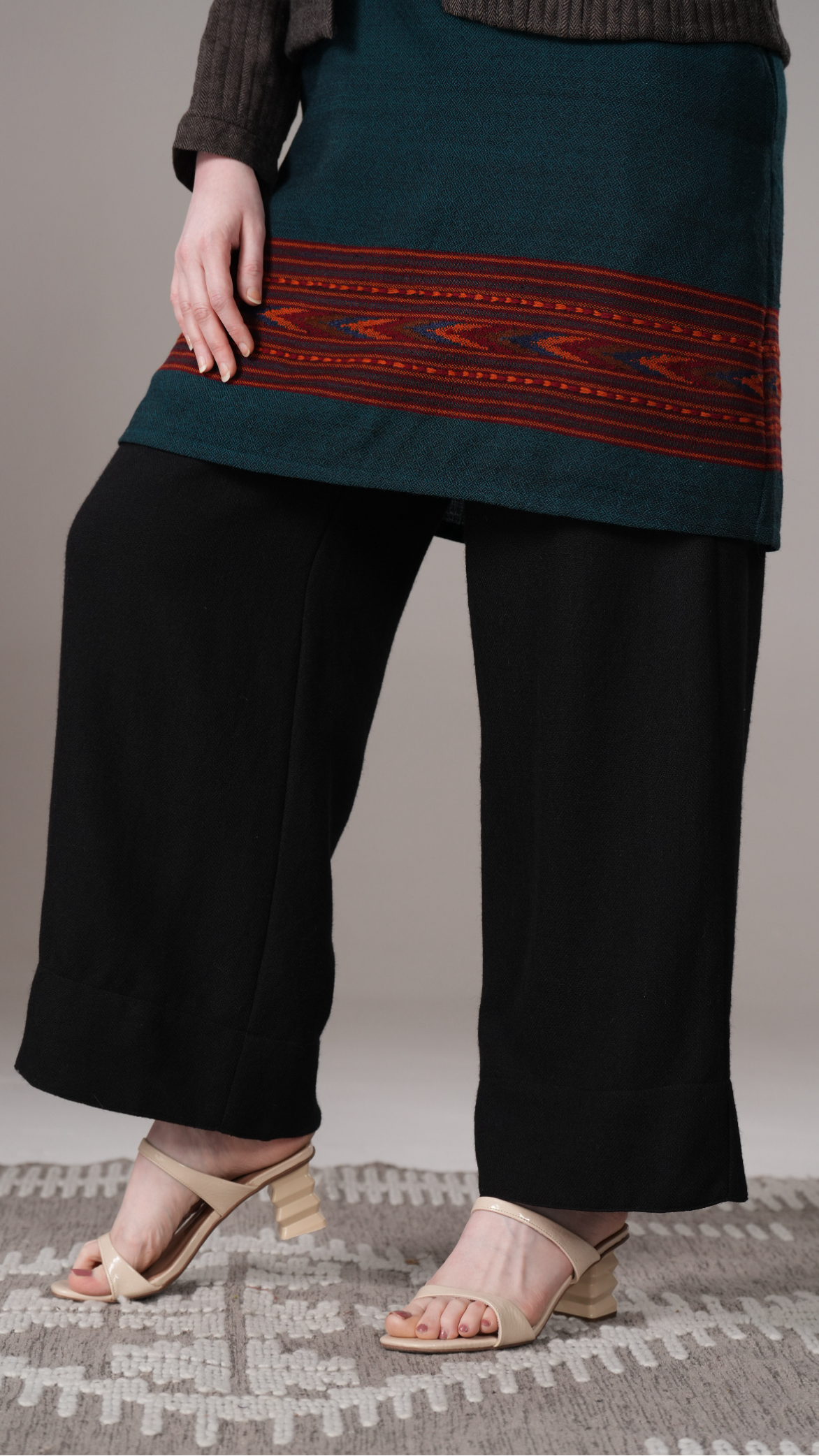 Person wearing a dark green top with red pattern and black pants on a neutral background