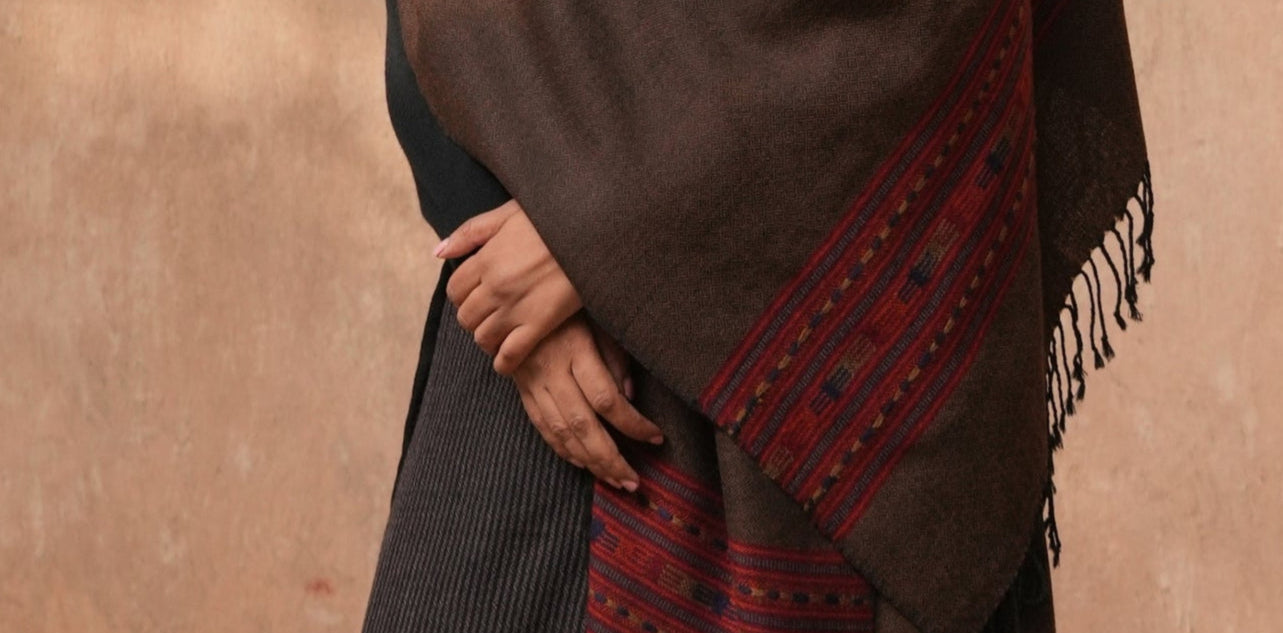 Person wearing a dark brown poncho with red patterns over a black dress, standing against a beige wall.