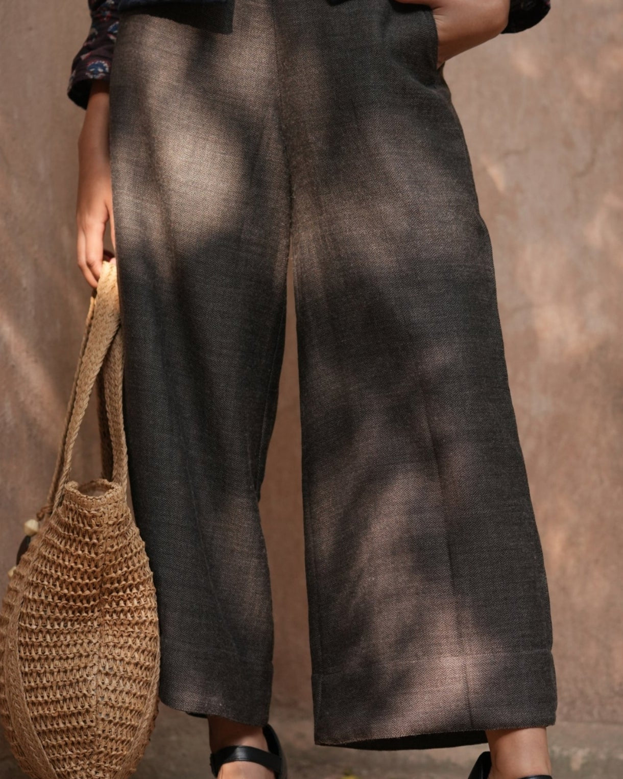 Person wearing wide-leg pants holding a woven bag against a textured wall.