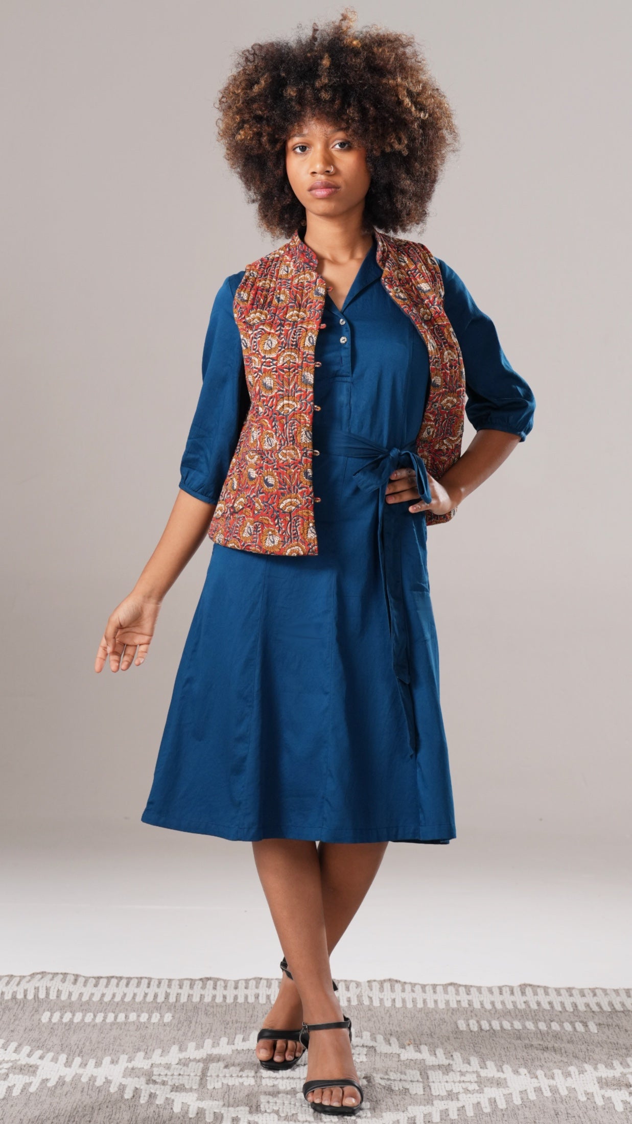 Woman wearing a blue dress with a patterned vest on a neutral background