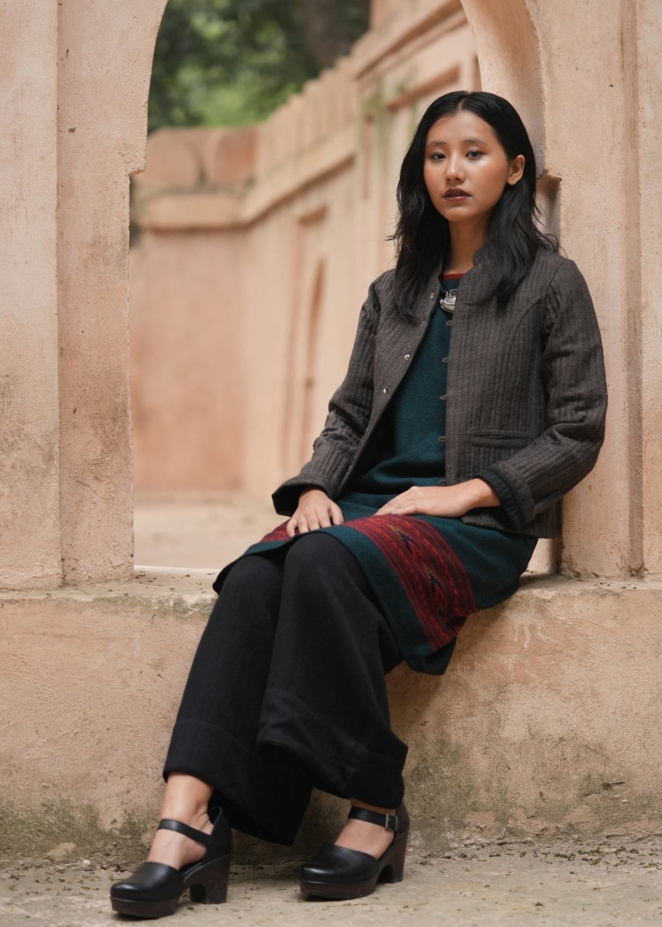 Woman sitting against a stone wall wearing a dark jacket and black pants.
